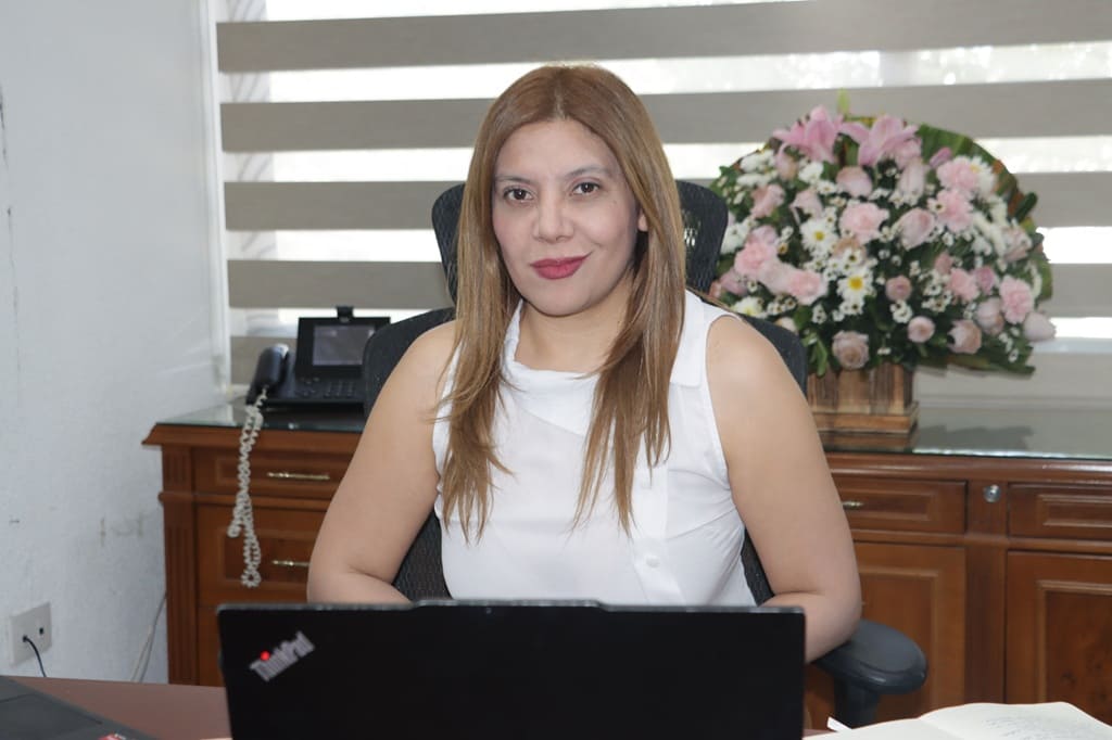 Mujer cabello castaño claro sentada frente a una computadora negra, vestida de blanco.