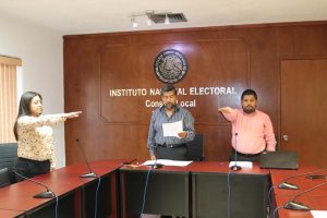 tres personas de pie, un hombre leyendo, mientras una mujer y otro hombre con el brazo derecho extendido al frente
