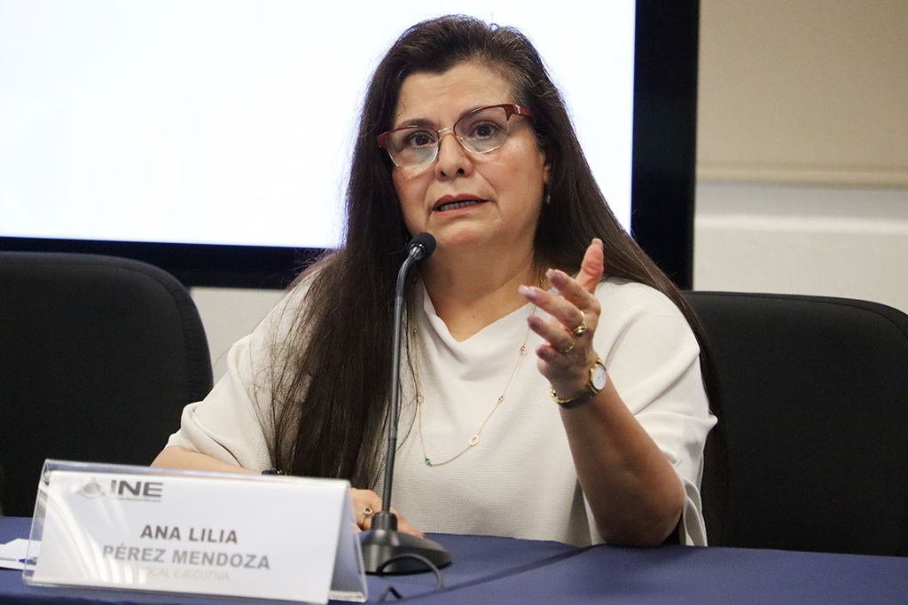 mujer de cabello largo lacio y con lentes sentada y hablando frente a un micrófono